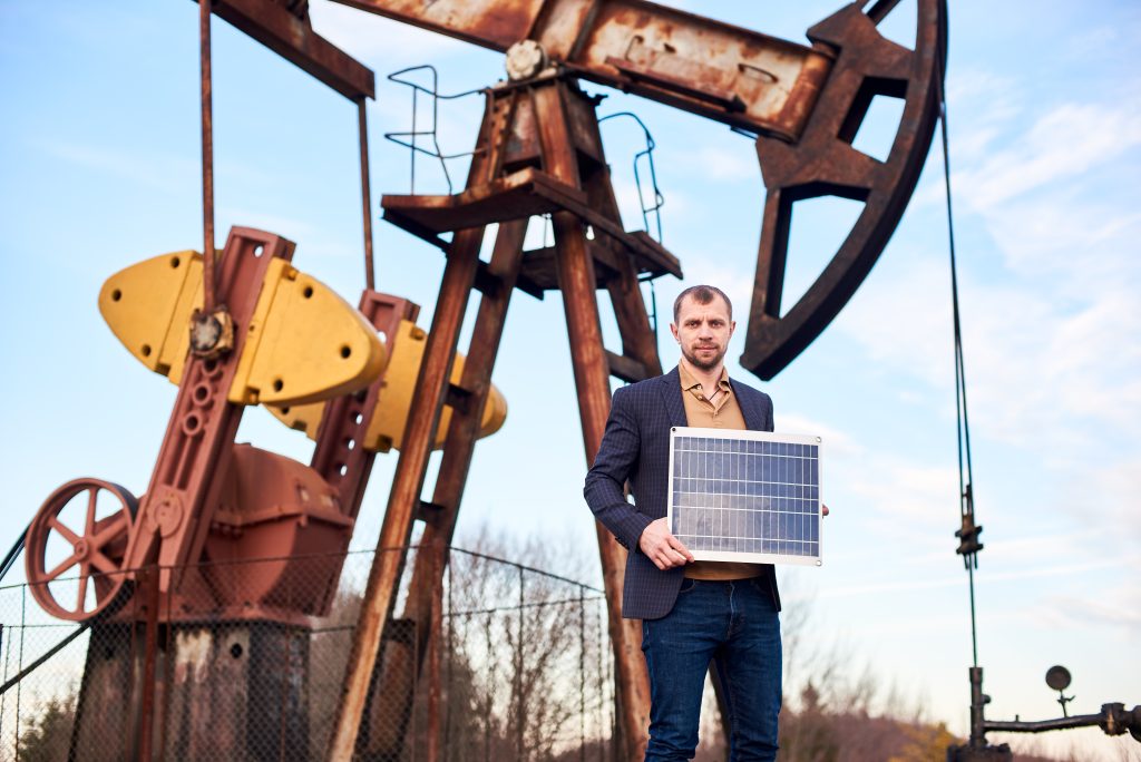 Businessman standing on an oilfield holding mini solar module next to an oil rig in canada